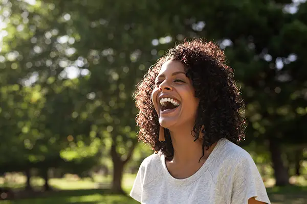 Happy woman laughing in a sunny park, showcasing the positive connection between oral care and general well-being.